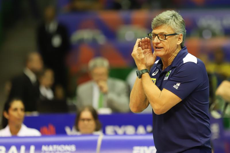 técnico da seleção brasileira de vôlei, homem com cabelos brancos e óculos preto, fazendo uma concha com a mão para falar. Ele está de uniforme azul marinho e usa um relógio preto