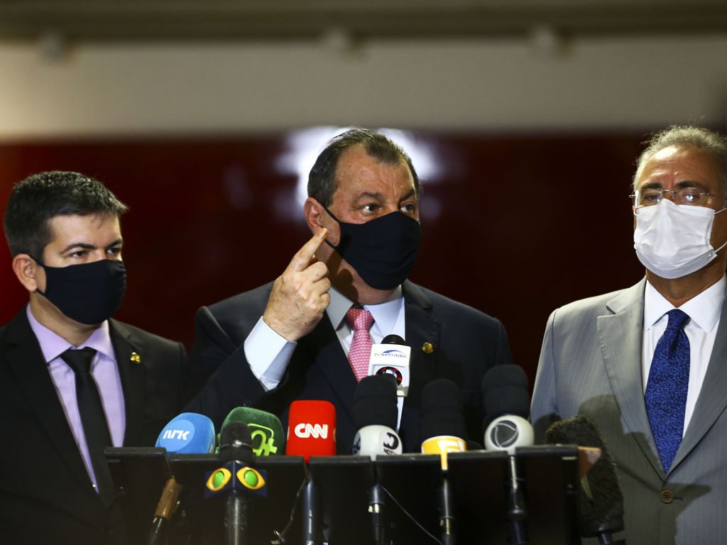Três homens brancos juntos falando em coletiva de imprensa, com microfones apoiados em um palanque. Os três usam máscara de proteção preta e o do meio está com o dedo levantado.