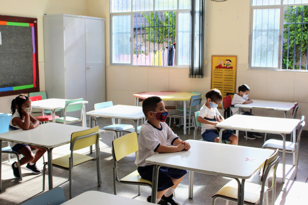 Quatro crianças sentadas na carteira escolar, uma distante da outra, assistem a uma aula em escola do Rio de Janeiro