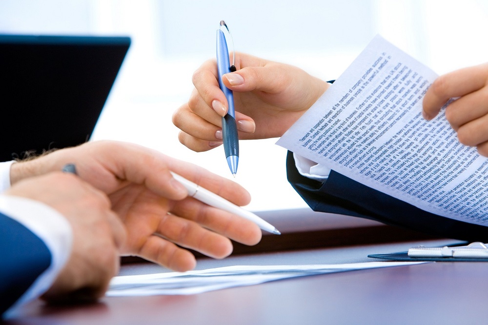 Imagem mostra as mãos de duas pessoas sobre uma mesa, sendo que ambas seguram uma caneta, mas apenas uma maneja um contrato
