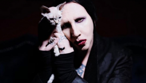 Cantor Marilyn Manson segurando um gato pequeno sem pelos. Ele tem o rosto bem branco, batom vermelho e usa roupa preta.