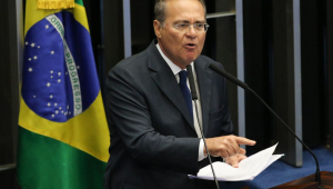 Senador Renan Calheiros discursando em tribuna, com um papel na mão e em frente a um microfone. Ele tem o dedo levantado e está falando. É um homem branco de óculos, com cabelos grisalhos e usa terno preto.