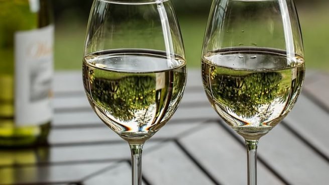 Duas taças com vinho branco servido até a metade apoiadas em uma mesa de madeira, que está em segundo plano