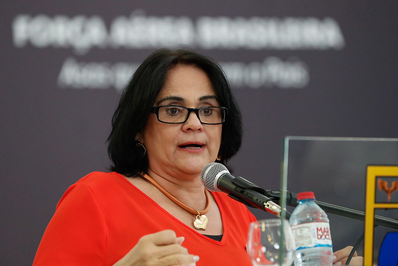 Ministra Damares Alves falando em microfone. Tem cabelos curtos pretos, usa óculos preto e vestido e acessórios vermelhos.