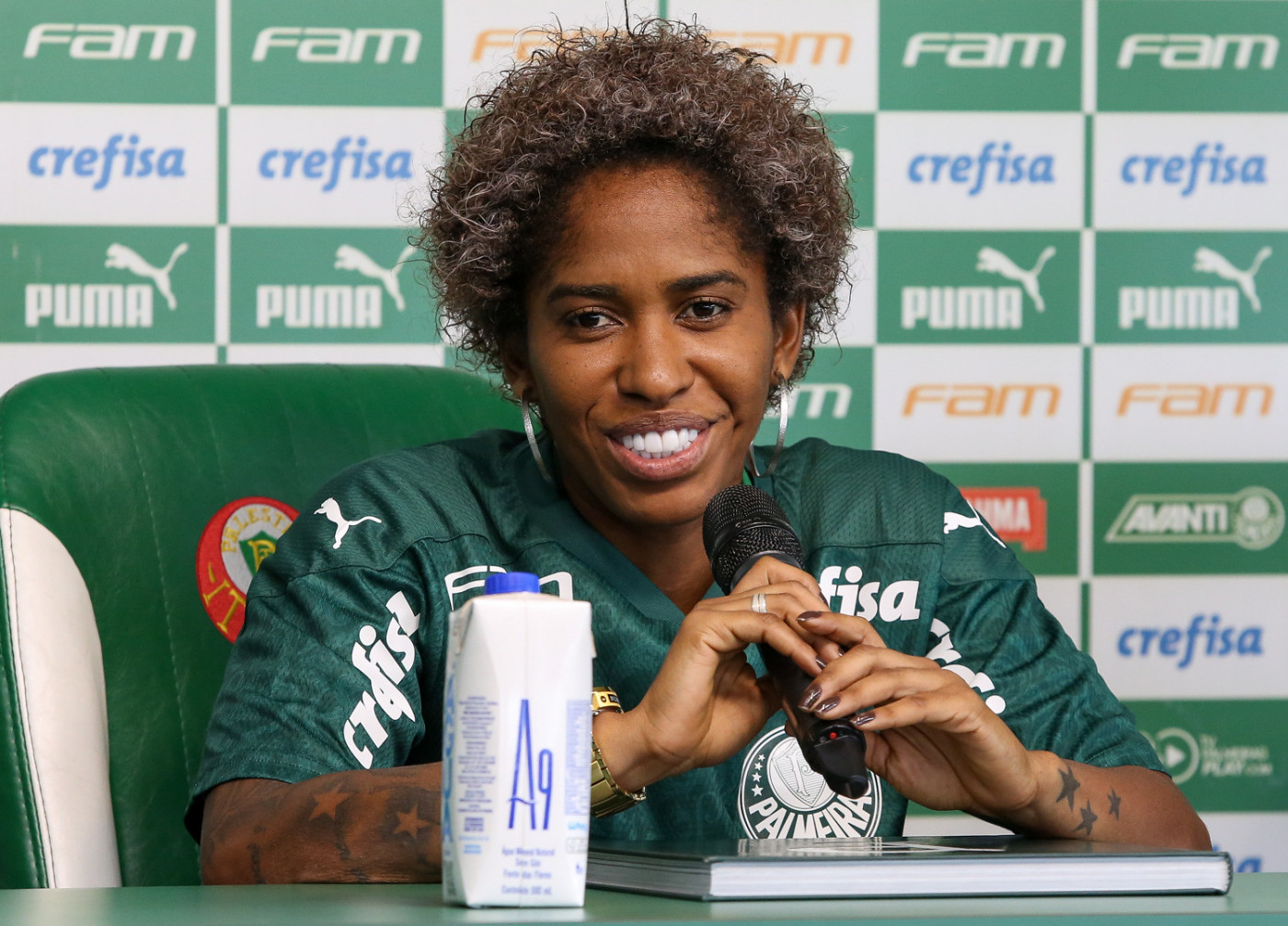 A atacante Chu, uma jogadora negra, beste a camisa do Palmeiras e, sentada, dá entrevista na sala de imprensa do clube