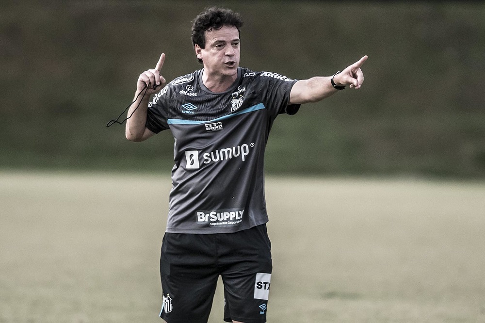 Com uma camisa cinza e um shorts preto do Santos, Fernando Diniz gesticula em um dos campos do CT Rei Pelé