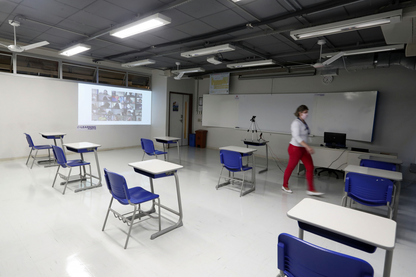 Sala de aulas em escola de São Paulo com retroprojetor, carteiras (separadas com distanciamento apropriado) e um monitor; mulher passa pela sala