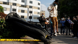 Enquanto um homem com chapéu, camisa e calça pula em frente à estátua de Gonzalo Jiménez de Quesada, outras pessoas tiram foto do monumento caído no chão