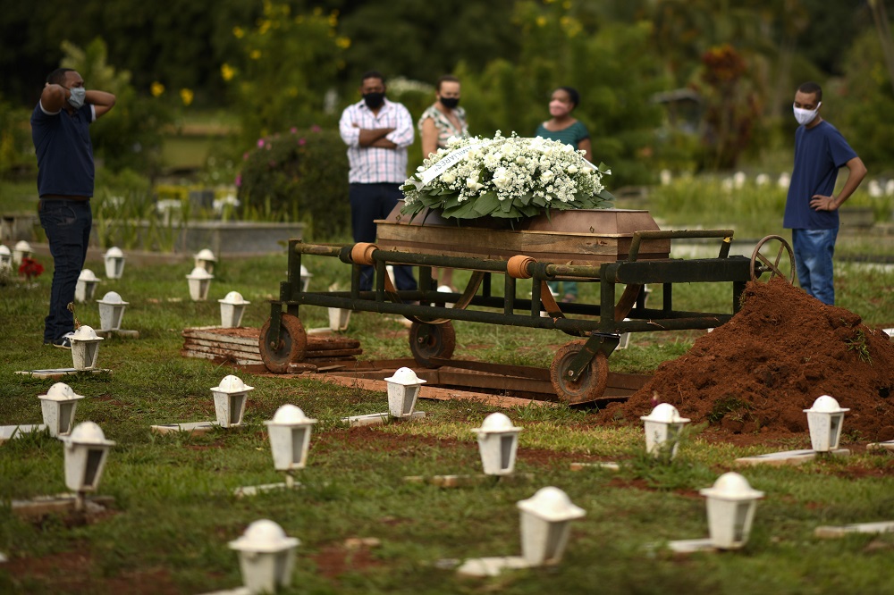 Cinco pessoas, sendo três homens e uma mulher, presenciam o enterro de um ente querido em um cemitério em São Paulo (três delas estão perto uma da outras, enquanto dois homens estão mais distantes)