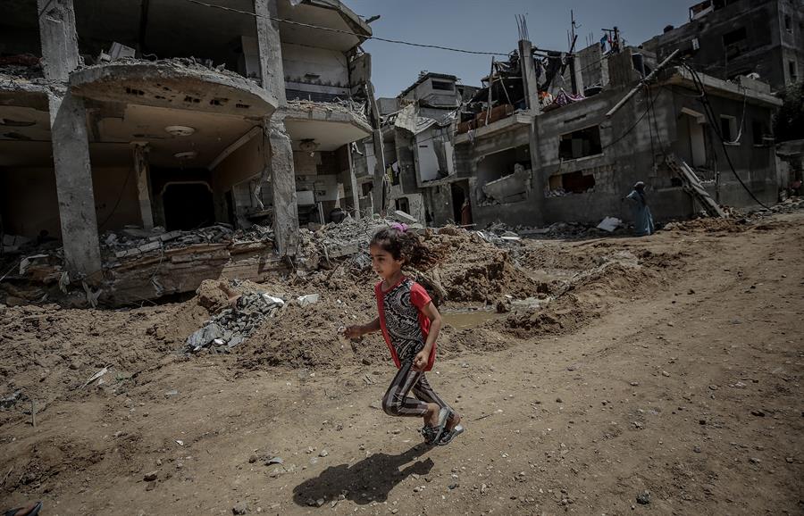 Imagem de criança correndo na Faixa de Gaza. Ao fundo, casas destruídas pela guerra