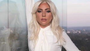 Lady Gaga com um braço levantado encostada em um prédio