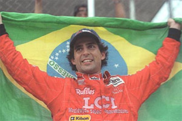 Andre Ribeiro de uniforme vermelho da Fórmula 1, boné preto, sorrindo e segurando uma bandeira do Brasil