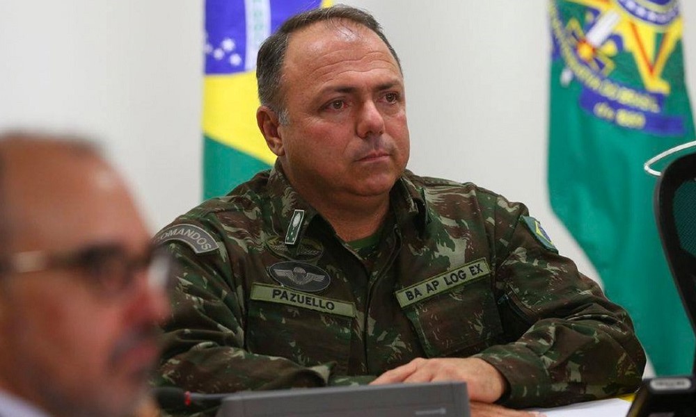 Vestido com roupa militar, o general Eduardo Pazuello aparece sentado atrás de uma mesa, com uma bandeira do Brasil atrás dele