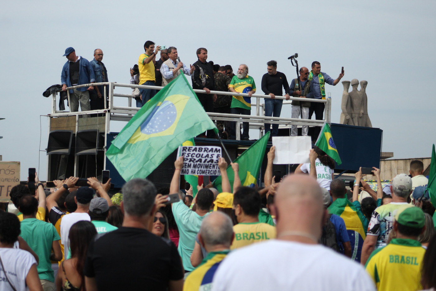 Jair Bolsonaro e outras pessoas em cima de um trio elétrico. Na foto, aparecem várias bandeira do Brasil e um público com camisetas e bandeiras