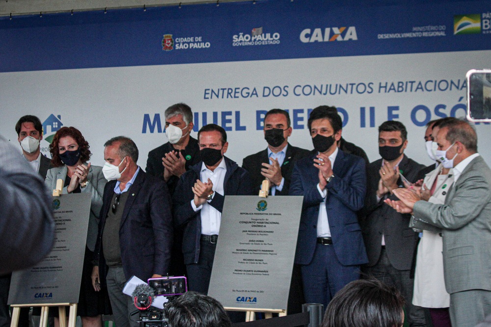 Doze políticos, entre eles Carla Zambelli (a única mulher, do lado esquerdo da imagem), Rogério Marinho (do lado direito da deputada), João Doria (no centro da foto) e Ricardo Nunes (perto de Doria, à direita), se apertam em palco durante inauguração de conjunto habitacional