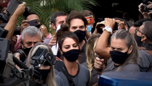 Monique Medeiros, mãe de henry borel, com multidão em volta. Ela usa blusa cinza, máscara de proteção preta e tem o cabelo preso em um rabo.