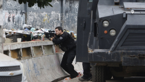 Ao lado do blindado, policial se esgueira sobre parapeito na favela e aponta o seu fuzil