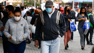 Dezenas de pessoas com máscaras de proteção e roupas de frio transitam por uma larga calçada em São Paulo
