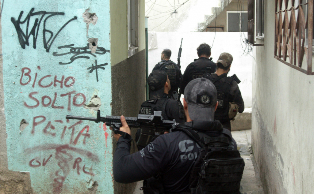 Grupo de policiais com coletes pretos e bonés empunhando armas dentro de uma favela