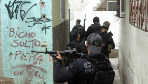 Grupo de policiais com coletes pretos e bonés empunhando armas dentro de uma favela