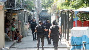 Em uma rua estreia da favela do Jacarezinho, cinco policiais civis andam pela rua, enquanto o caminhão blindado da polícia anda em direção a eles; três crianças e duas mulheres assistem a tudo da calçada
