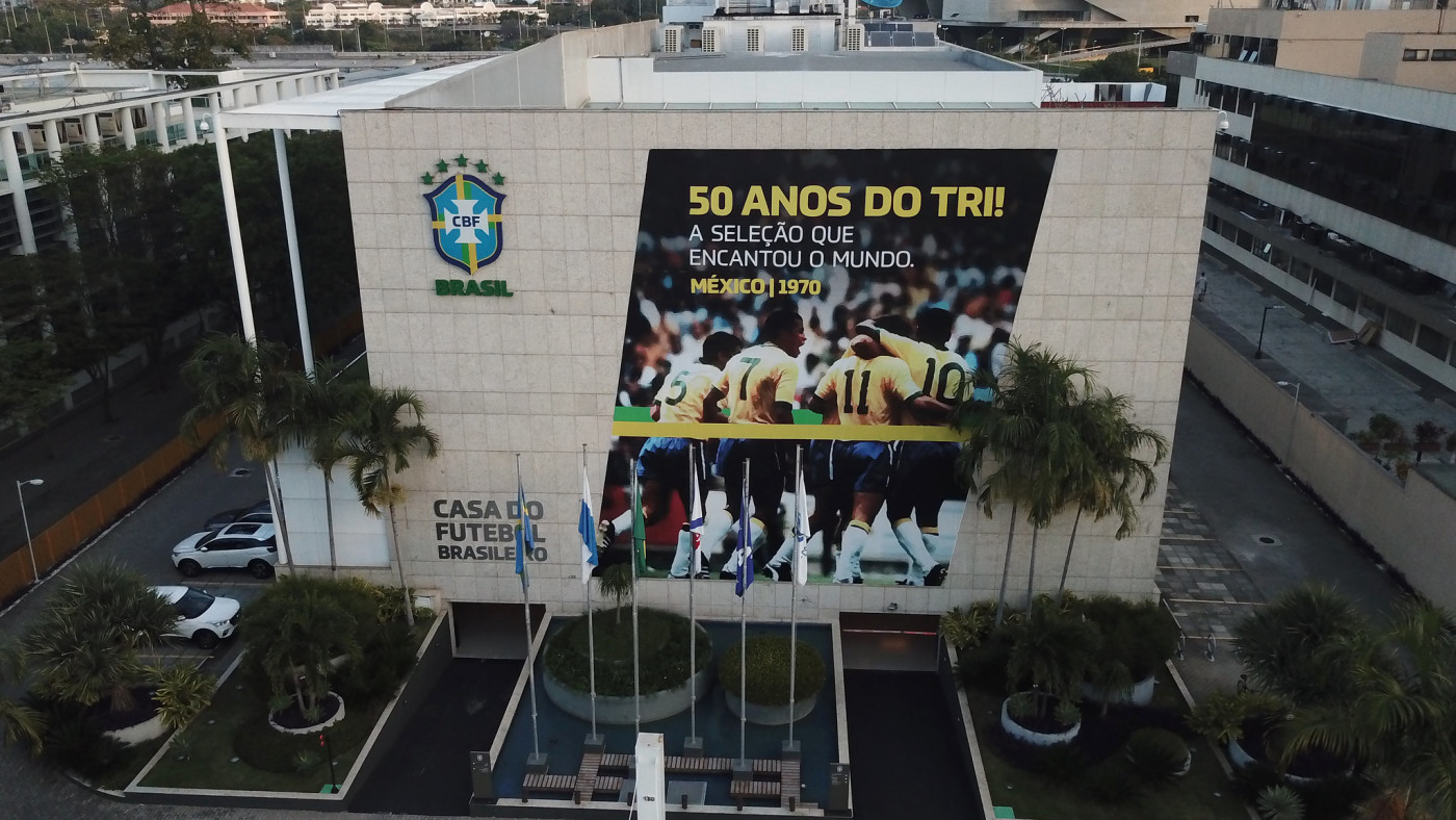 Sede da Confederação Brasileira de Futebol (CBF) na Barra da Tijuca. Prédio com um banner escrito '50 anos do Tri' com jogadores comemorando. O prédio é de cor clara e tem algumas árvores na frente e o símbolo da CBF