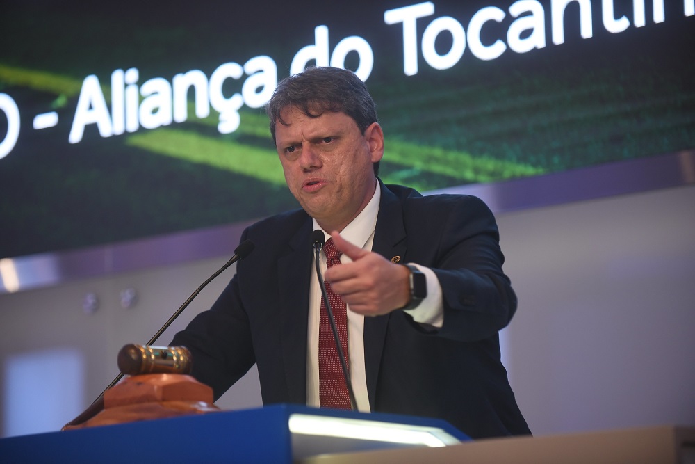 De terno preto, camisa branca e gravata vermelha, o ministro Tarcísio de Freitas fala de cima de um palco onde se vê um martelo de leilão