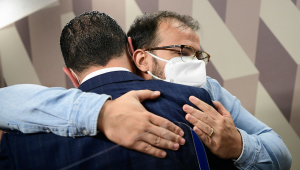Dois homens se abraçando usando máscaras de proteção. Um está de costas para a câmera e o outro o abraça de frente