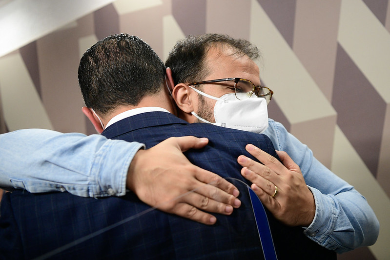 Dois homens se abraçando usando máscaras de proteção. Um está de costas para a câmera e o outro o abraça de frente