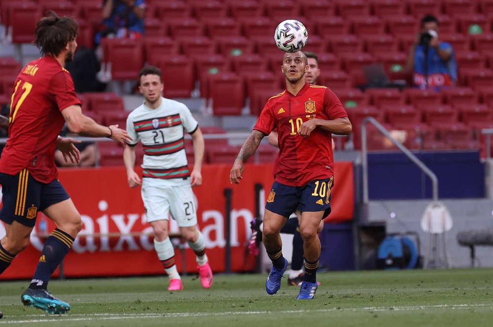 Vestido com a camisa vermelha e o shorts azul da seleção espanhola, Thiago Alcântara amortece a bola na cabeça durante jogo contra Portugal; um companheiro está perto dele e um rival o observa de trás