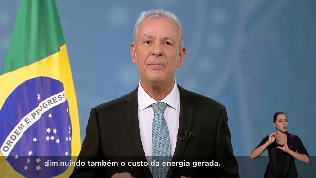 Ministro Bento Albuquerque, homem de terno preto, camisa branca e gravata azul, falando na televisão. Ao lado, uma bandeira do Brasil sem estar hasteada