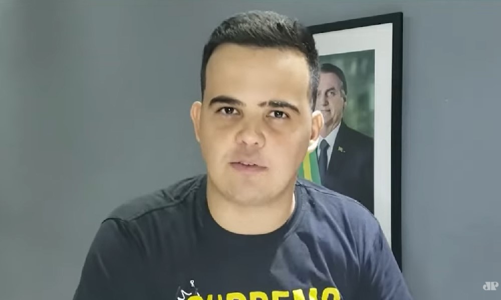 De camiseta cinza escura, em um cômodo com paredes cinzas e um quadro de Jair Bolsonaro pendurado, o deputado Cano Junio Amaral (homem branco, cabelo preto curto e ondulado, 33 anos) grava depoimento para a Jovem Pan