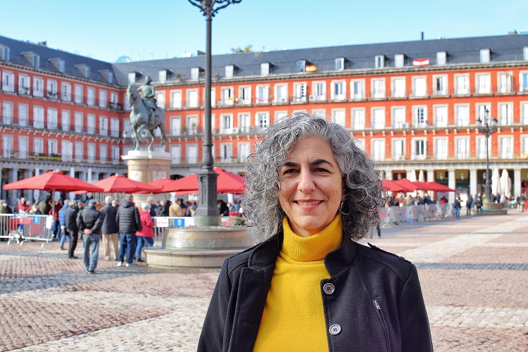 Mulher olhando para foto com uma blusa de lã amarela e um casaco preto. Ela tem cabelos grisalhos e está em um cenário que é a Plaza Mayor, com uma estátua e prédios baixos em volta