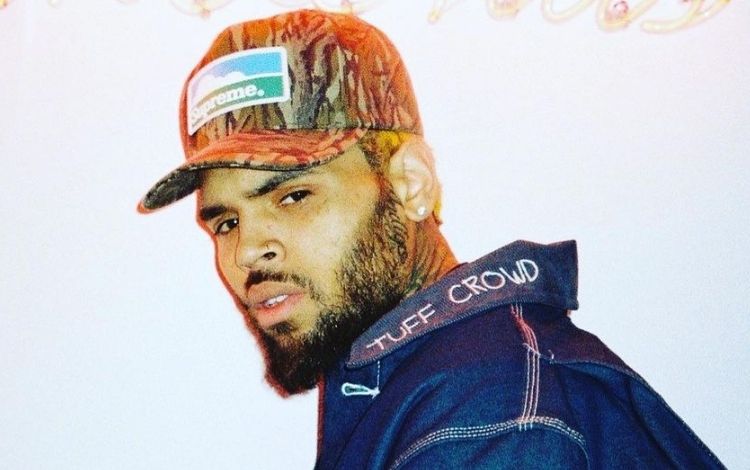 Chris Brown com um boné e jaqueta de lado, mas olhando sério para frente