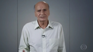 Drauzio Varella, idoso careca de camisa branca de botões, fala em frente à câmera