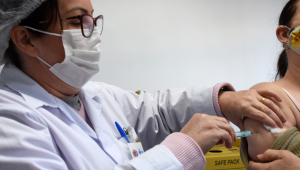 Enfermeira (que usa óculos e máscara branca) aplica vacina no braço esquerdo de uma mulher, que aparece apenas parcialmente na imagem
