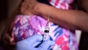 Ao fundo, mulher grávida repousa a mão sobre a barriga enquanto um profissional da saúde mostra vacina contra a Covid-19