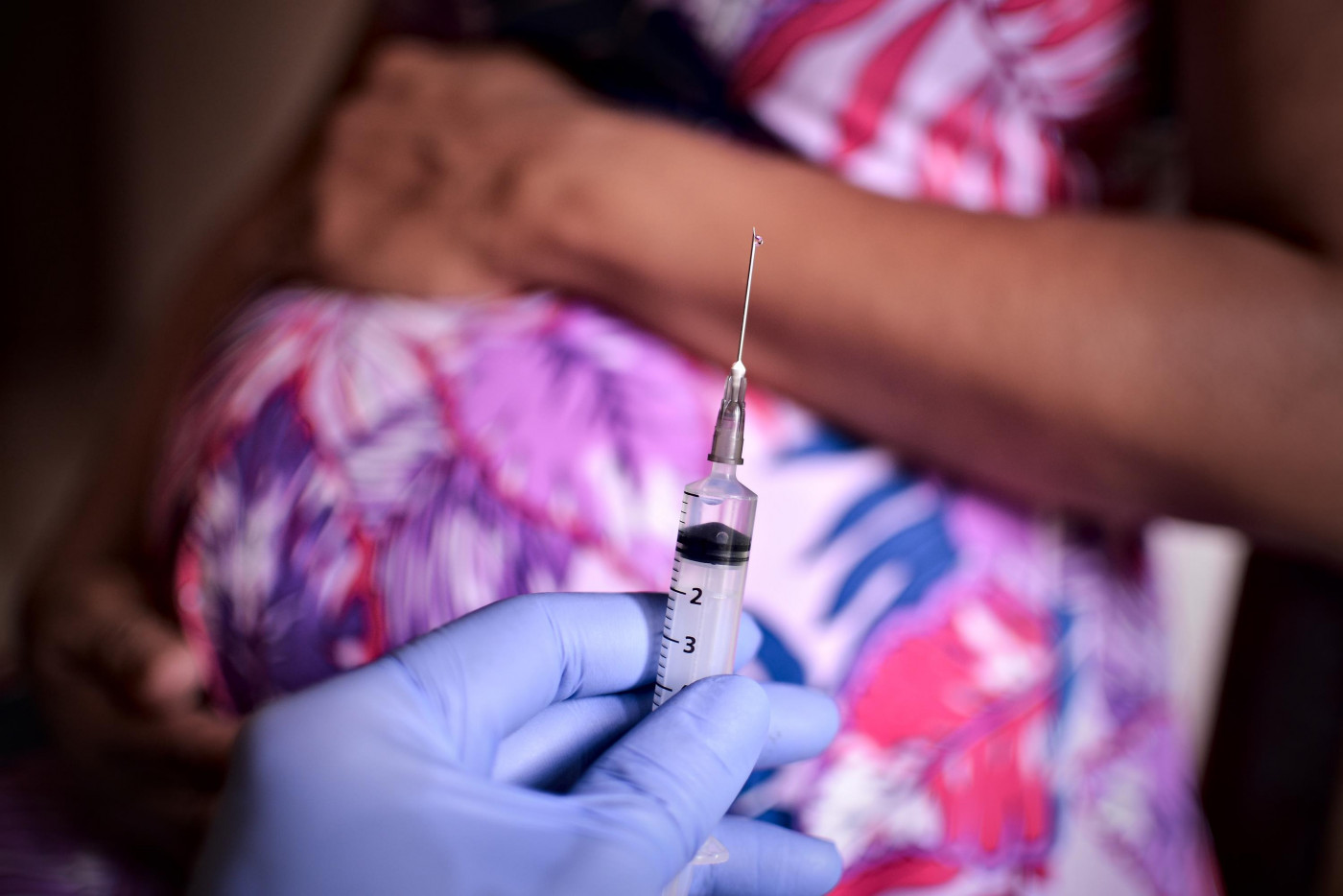 Ao fundo, mulher grávida repousa a mão sobre a barriga enquanto um profissional da saúde mostra vacina contra a Covid-19