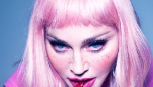 Madonna com uma peruca rosa channel, olhos azuis com sombra preta e as bochechas rosadas. Ela está com a boca rosada e morde uma bala