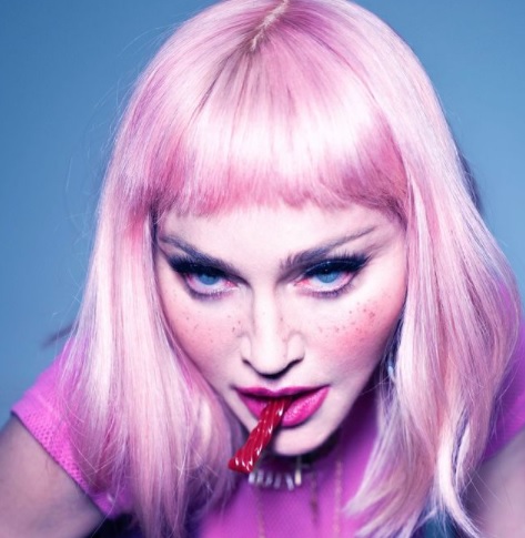 Madonna com uma peruca rosa channel, olhos azuis com sombra preta e as bochechas rosadas. Ela está com a boca rosada e morde uma bala