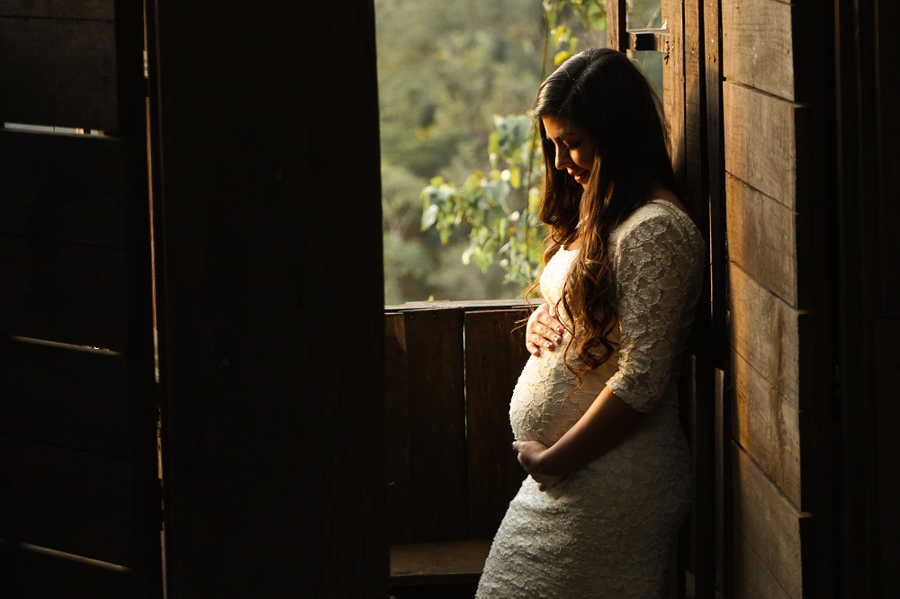 Em um cômodo escuro, com uma janela aberta (por onde se vê vegetação do lado de fora), grávida passa a mão na barriga