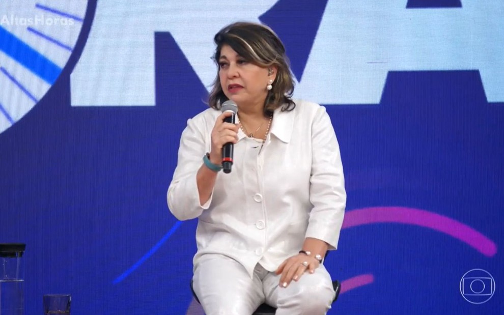 Roberta Miranda com uma camisa e calça branca com um microfone na mão, sentada em um banco, no programa Altas Horas. Mulher branca, de cabelos castanhos com mechas loiras channel