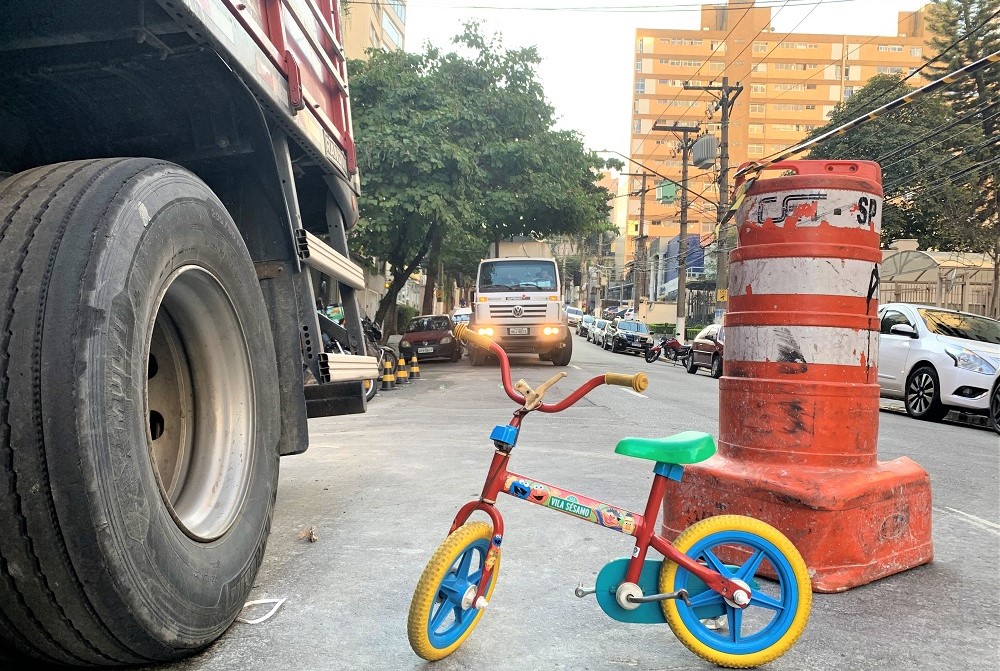 Pequena bicicleta multicolorida está parada na rua bem perto de um cone da CET e de um caminhão