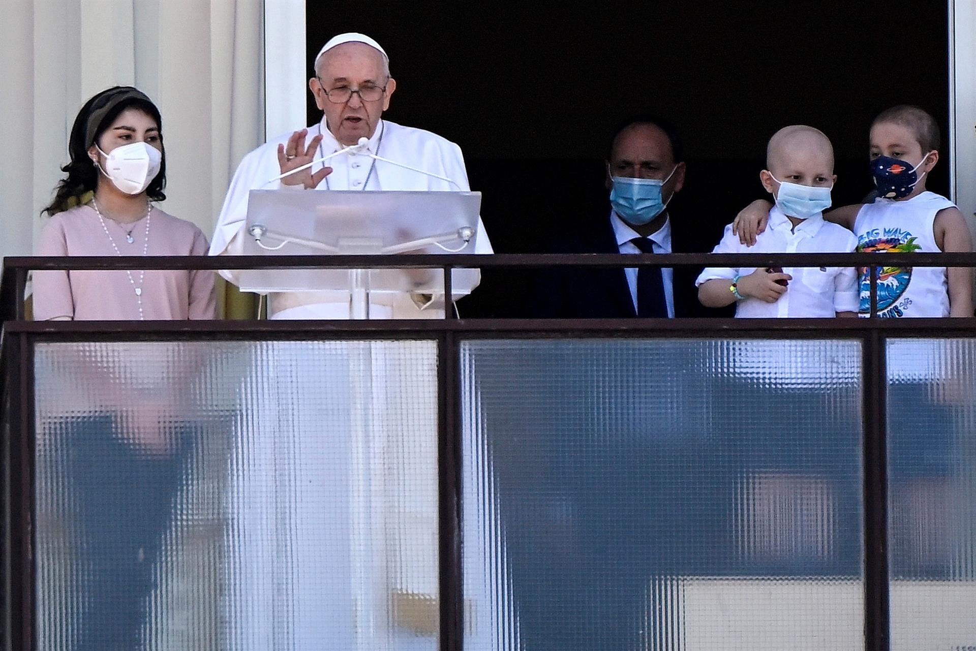 Papa Francisco lendo um texto em varanda de um hospital. Ao lado, duas crianças usando máscaras e um homem de terno atrás delas