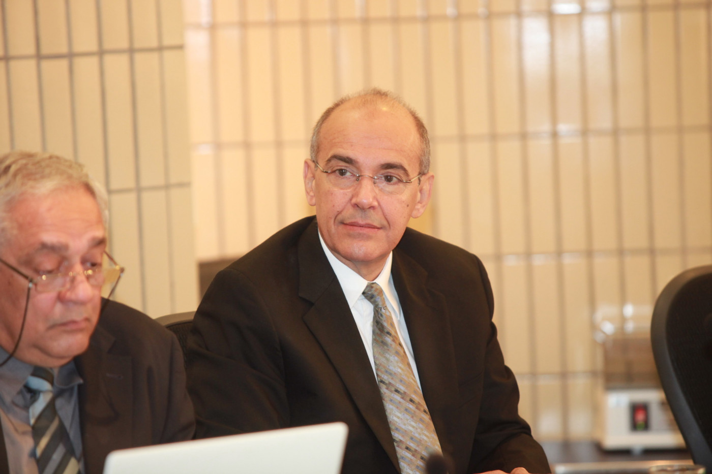 Homem careca usando terno e óculos de grau olhando para a câmera ao lado de outro homem em uma reunião