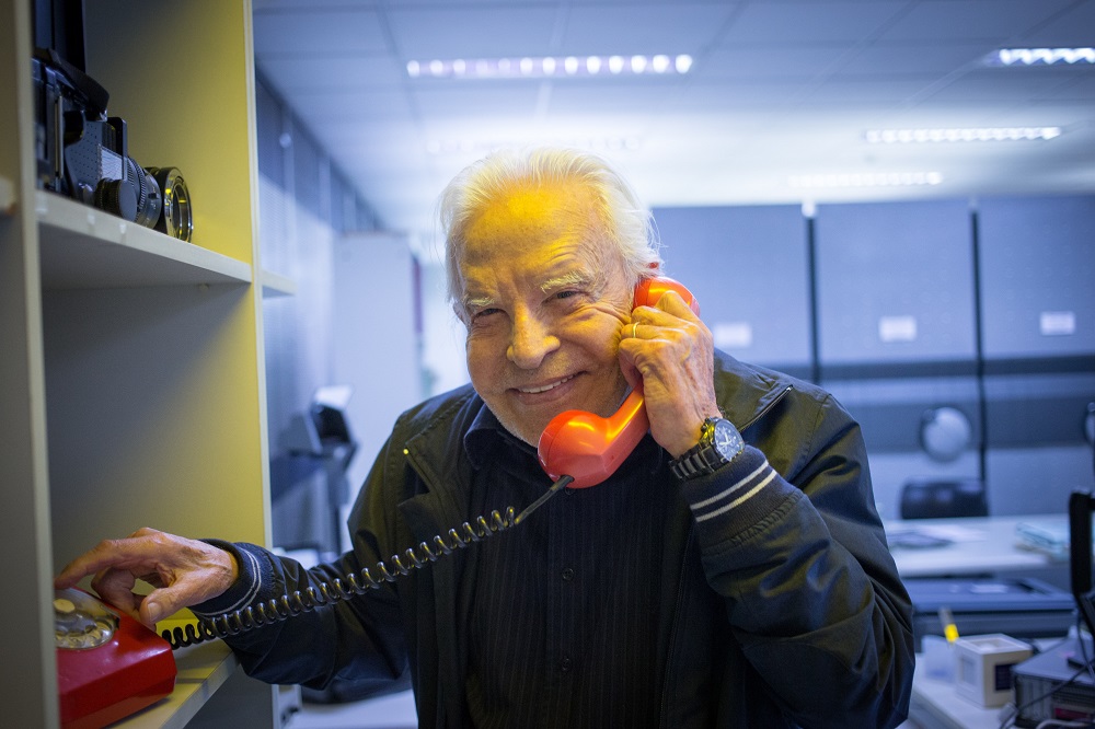 O jornalista Cid Moreira, então com 89 anos, posa sorrindo segurando um telefone rente à orelha