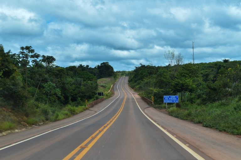 Foto de uma rodovia com uma faixa amarela no meio. Ao fundo céu azul com nuvens e aos lados árvores
