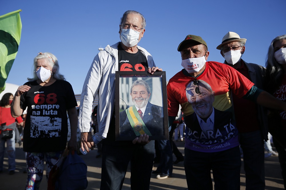 Com uma camiseta preta, calça jeans e jaqueta cinza clara, José Dirceu segura quadro de Lula ao lado de militantes petistas durante protesto contra Jair Bolsonaro em Brasília