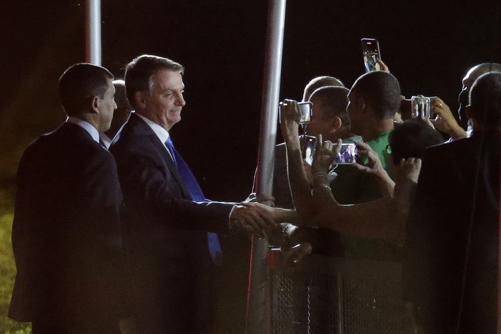 De terno azul marinho, Jair Bolsonaro conversa com apoiadores, que estão no cercadinho do Palácio do Alvorada; percebe-se que está de noite