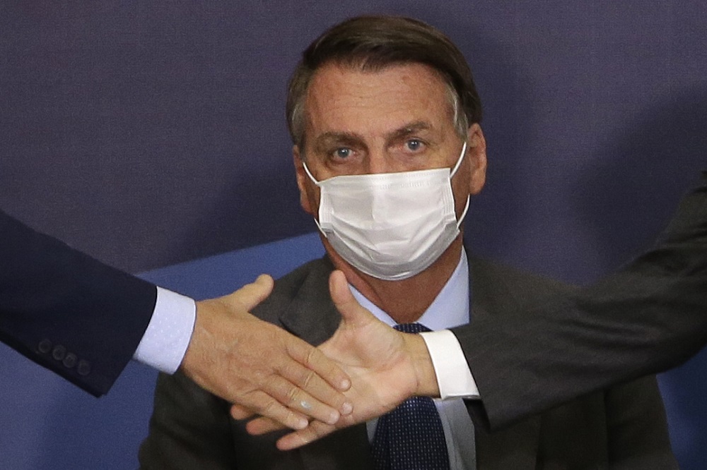 De traje social e máscara, presidente Jair Bolsonaro tem olhar perdido enquanto duas pessoas fazer cumprimento com as mãos à sua frente